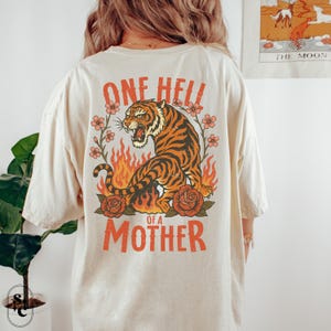 Puede incluir: Camiseta color crema con un gr&aacute;fico de tigre, llamas, rosas y el texto "ONE HELL OF A MOTHER" en naranja. El tigre es naranja y negro, con una expresi&oacute;n feroz. Acentos florales rodean al tigre.