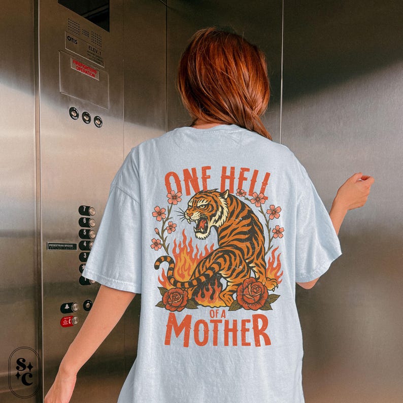 Puede incluir: Camiseta azul claro con un dise&ntilde;o de tigre. El dise&ntilde;o presenta un tigre rugiente rodeado de llamas, rosas y el texto "ONE HELL OF A MOTHER" en naranja. La camiseta es de corte holgado.