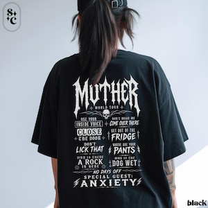 May include: A black short-sleeved t-shirt featuring white graphic text on the back. The design includes the word MUTHER, WORLD TOUR, and phrases like USE YOUR INSIDE VOICE, CLOSE THE DOOR, DON'T LICK THAT, GET OUT OF THE FRIDGE, and ANXIETY.