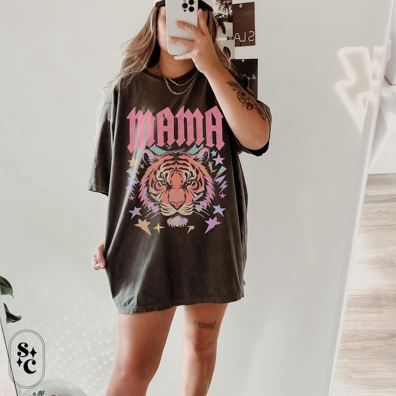May include: Dark grey t-shirt featuring the word "MAMA" in pink block letters above a colourful tiger face graphic. The tiger has pink, orange, and yellow stripes, with stars and other shapes around it. The shirt is a casual, oversized style.