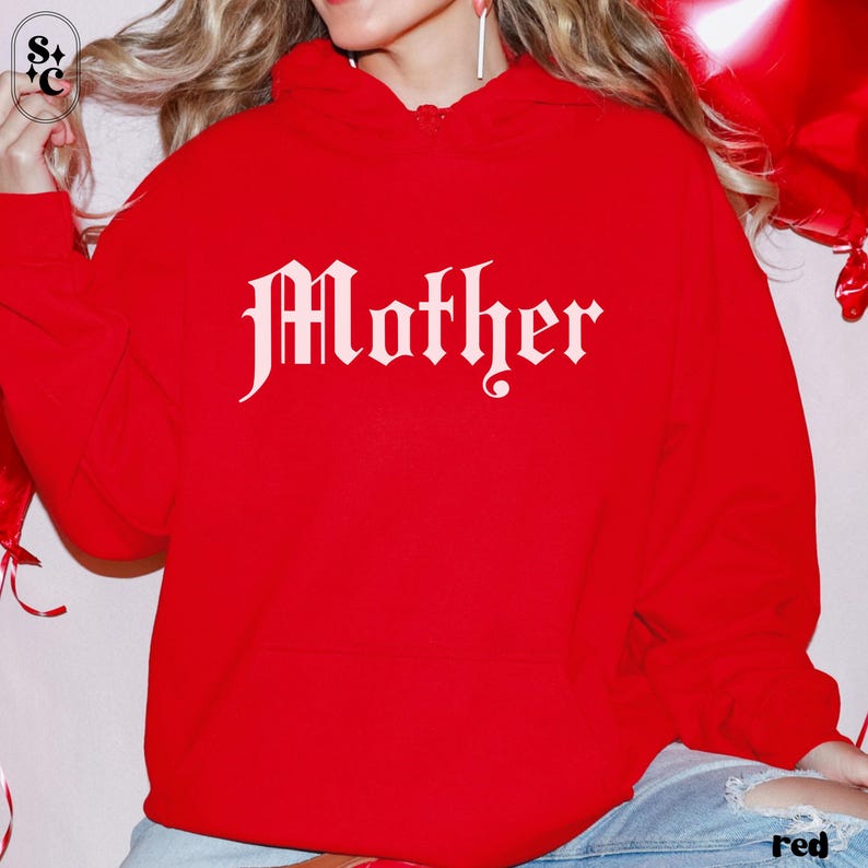 May include: A red hoodie with the word "Mother" in white gothic lettering. The hoodie is a solid red color and has a drawstring hood. The image is of a person wearing the hoodie.