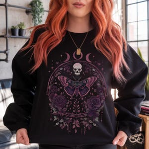 Puede incluir: Sudadera negra con un diseño circular que presenta una calavera, medias lunas y una gran polilla. La polilla tiene alas moradas y rosas, rodeadas de rosas y follaje en tonos morados y rosas.