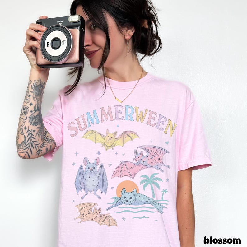 May include: A light pink t-shirt with the word "SUMMERWEEN" in pastel colors and illustrations of bats, a sun, and palm trees. The shirt has a crew neck and short sleeves.