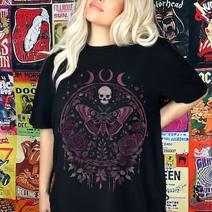 K&ouml;nnte beinhalten: Schwarzes T-Shirt mit einem grafischen Druck eines Sch&auml;dels, eines Schmetterlings und Blumen in Rosa- und Lilat&ouml;nen. Die Grafik ist von einem Kreis mit drei Mondsicheln umgeben.