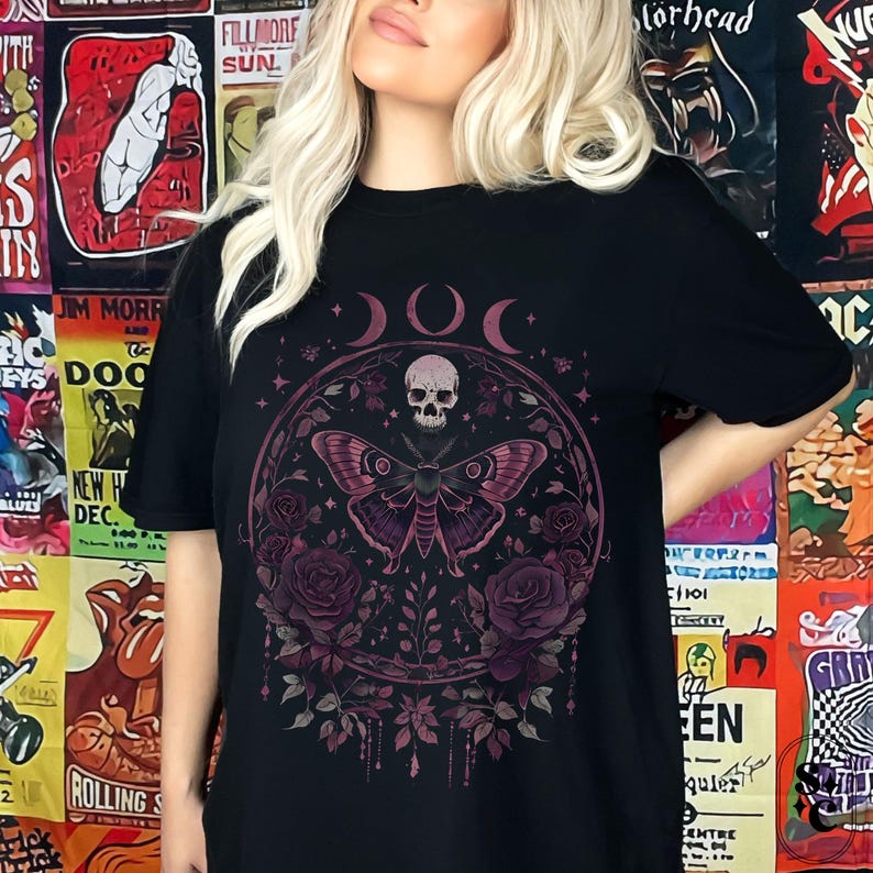 K&ouml;nnte beinhalten: Schwarzes T-Shirt mit einem grafischen Druck eines Sch&auml;dels, eines Schmetterlings und Blumen in Rosa- und Lilat&ouml;nen. Die Grafik ist von einem Kreis mit drei Mondsicheln umgeben.