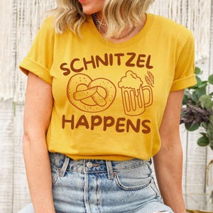 May include: Mustard yellow t-shirt with the phrase "SCHNITZEL HAPPENS" in brown lettering. The design includes a pretzel, a beer stein, and wheat stalks. The model is wearing blue jeans.