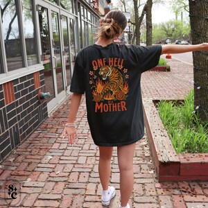 Puede incluir: Camiseta negra con un dise&ntilde;o de tigre y el texto "ONE HELL of a MOTHER" en naranja. El tigre est&aacute; rodeado de llamas y flores. La camiseta la lleva una persona caminando por un camino de ladrillos.