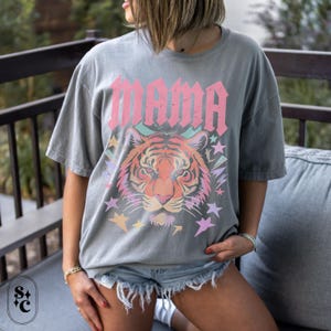May include: Grey t-shirt featuring the word "MAMA" in pink, above a tiger face with colourful stars. The shirt is worn by a person wearing denim shorts. The t-shirt has short sleeves and a relaxed fit.