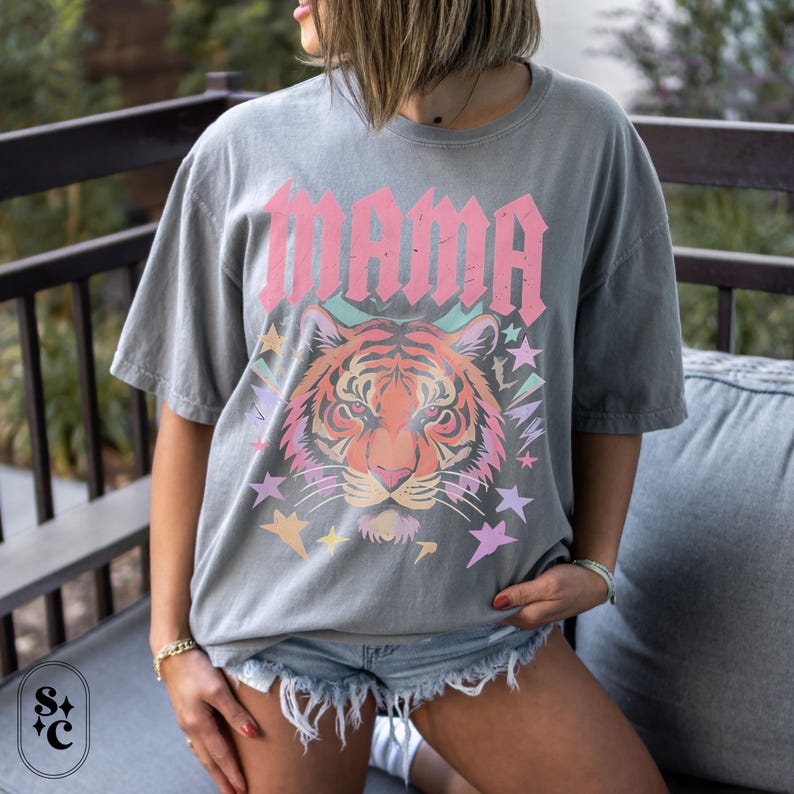 May include: Grey t-shirt featuring the word "MAMA" in pink, above a tiger face with colourful stars. The shirt is worn by a person wearing denim shorts. The t-shirt has short sleeves and a relaxed fit.