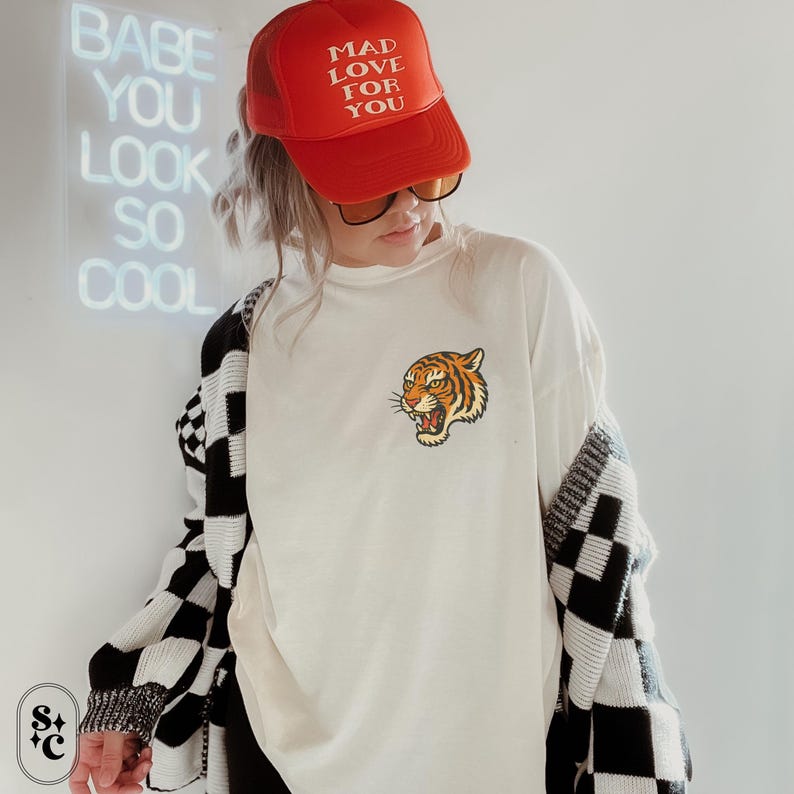Puede incluir: Una camiseta blanca con un gr&aacute;fico de cabeza de tigre, combinada con una gorra de camionero roja que dice "MAD LOVE FOR YOU". El atuendo incluye una rebeca a cuadros en blanco y negro.