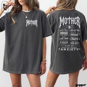 May include: A charcoal gray short-sleeve t-shirt featuring the word MOTHER in a white gothic font on the front chest. The back displays the same MOTHER text above a list of phrases including USE YOUR INSIDE VOICE, CLOSE THE DOOR, and ANXIETY.