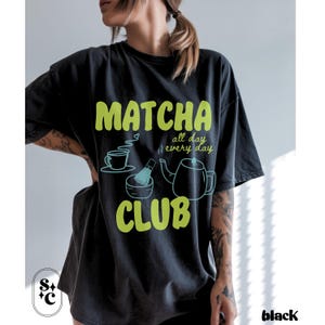 Puede incluir: Camiseta negra con tem&aacute;tica de matcha. La camiseta presenta las palabras "Matcha Club" en verde brillante, junto con ilustraciones de una taza de t&eacute;, un batidor y una tetera. Tambi&eacute;n se incluye el texto "all day every day".