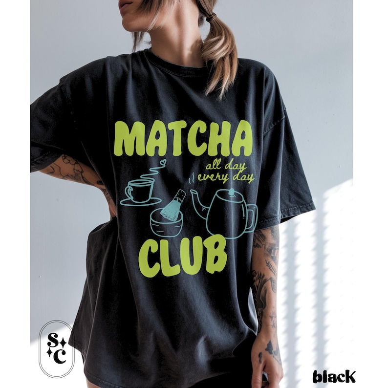 Puede incluir: Camiseta negra con tem&aacute;tica de matcha. La camiseta presenta las palabras "Matcha Club" en verde brillante, junto con ilustraciones de una taza de t&eacute;, un batidor y una tetera. Tambi&eacute;n se incluye el texto "all day every day".
