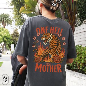 Puede incluir: Camiseta gris oscuro con un dise&ntilde;o de tigre. El tigre naranja y negro est&aacute; rodeado de llamas, flores y el texto "ONE HELL OF A MOTHER" en naranja. La camiseta es de corte holgado.