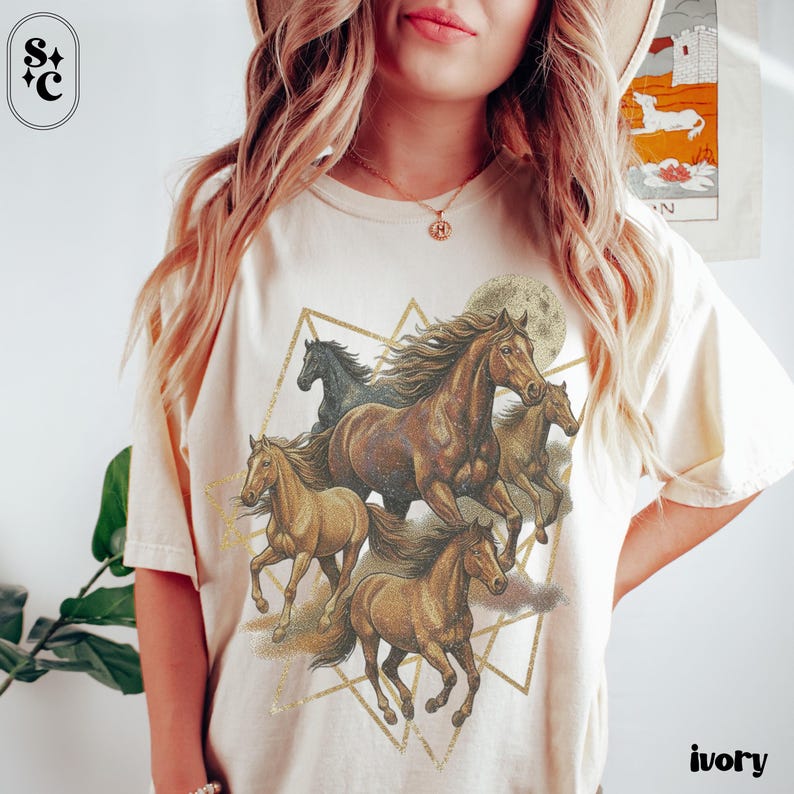 May include: Ivory-coloured t-shirt with a graphic of running horses, a full moon, and geometric shapes. The horses are in shades of brown and gold. The shirt has the word "ivory" printed on the bottom right.
