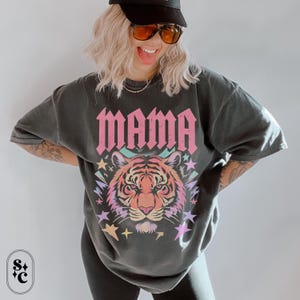 May include: Dark grey t-shirt featuring the word "MAMA" in pink block letters above a colourful tiger face design. The tiger has orange, pink, and yellow hues, surrounded by stars and pastel colours. The shirt has a relaxed fit.