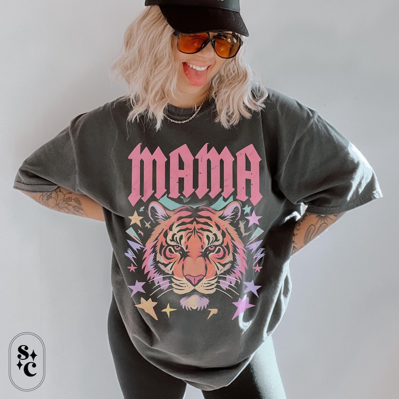 May include: Dark grey t-shirt featuring the word "MAMA" in pink block letters above a colourful tiger face design. The tiger has orange, pink, and yellow hues, surrounded by stars and pastel colours. The shirt has a relaxed fit.