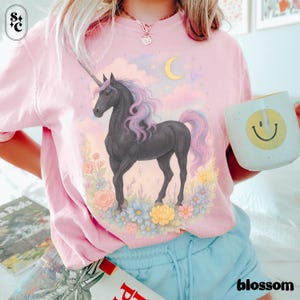 Peut inclure: T-shirt rose clair avec un motif de licorne noire, crini&egrave;re et queue violettes et roses, entour&eacute;e de fleurs et d'un croissant de lune. Le mot "blossom" est imprim&eacute; en blanc en bas &agrave; droite.