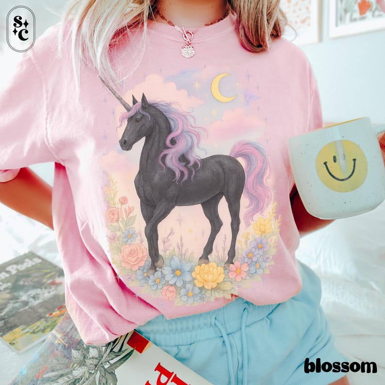 Peut inclure: T-shirt rose clair avec un motif de licorne noire, crini&egrave;re et queue violettes et roses, entour&eacute;e de fleurs et d'un croissant de lune. Le mot "blossom" est imprim&eacute; en blanc en bas &agrave; droite.