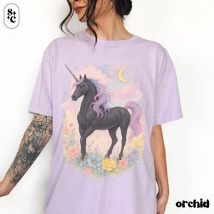 Peut inclure: T-shirt lilas avec un motif de licorne noire, crini&egrave;re et queue roses et mauves, entour&eacute;e de fleurs. Un croissant de lune et des nuages sont en arri&egrave;re-plan. Le mot "orchid" est imprim&eacute; en noir.