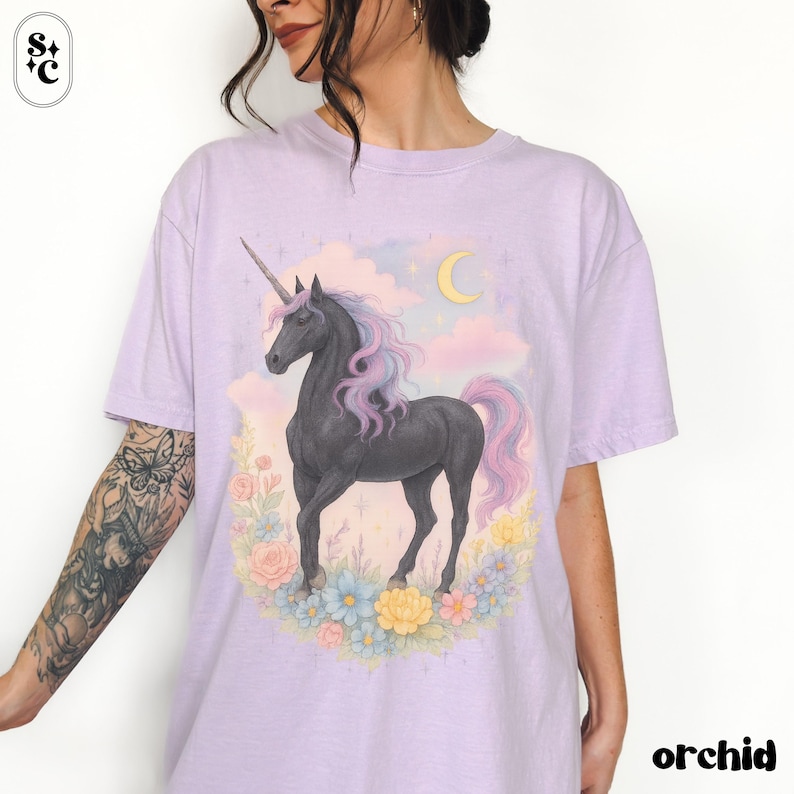 Peut inclure: T-shirt lilas avec un motif de licorne noire, crini&egrave;re et queue roses et mauves, entour&eacute;e de fleurs. Un croissant de lune et des nuages sont en arri&egrave;re-plan. Le mot "orchid" est imprim&eacute; en noir.