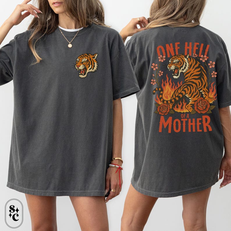 Puede incluir: Camiseta gris oscuro con dise&ntilde;o de tigre. La parte delantera tiene una peque&ntilde;a cabeza de tigre, mientras que la parte trasera muestra un tigre en llamas, flores y el texto "ONE HELL OF A MOTHER".