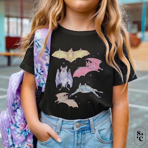 May include: A black short-sleeve t-shirt featuring a graphic print of five bats in various poses and colors, including yellow, pink, and blue, accented with small stars and hearts. The shirt is paired with light blue denim jeans and a floral backpack.