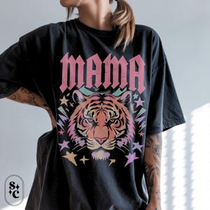 May include: Black t-shirt featuring the word "MAMA" in pink gothic font above a tiger illustration. The tiger is in shades of orange and pink, with pastel stars and lightning bolts.