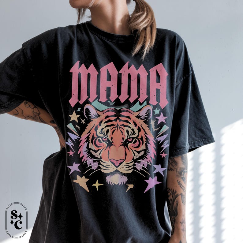 May include: Black t-shirt featuring the word "MAMA" in pink gothic font above a tiger illustration. The tiger is in shades of orange and pink, with pastel stars and lightning bolts.