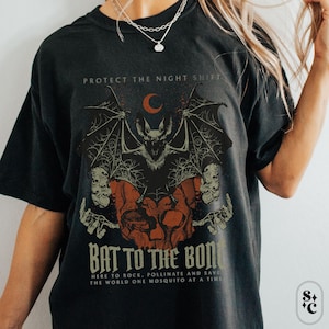 Op de afbeelding: Donkergrijs t-shirt met een vleermuis- en schedelafbeelding, een halve maan en de tekst "PROTECT THE NIGHT SHIFT" en "BAT TO THE BONE". Het shirt heeft ook de tekst "HERE TO ROCK, POLLINATE AND SAVE THE WORLD ONE MOSQUITO AT A TIME."