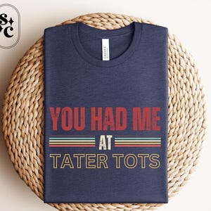 May include: A navy blue t-shirt with the text "You Had Me At Tater Tots" in a vintage style font. The text is in red, white, and blue.