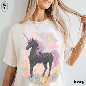 Peut inclure: T-shirt ivoire avec un motif de licorne noire. La licorne a une crini&egrave;re et une queue roses et violettes, sur fond de ciel pastel avec un croissant de lune. Une couronne florale de fleurs pastel entoure la licorne. Le mot "ivoire" est imprim&eacute; en bas &agrave; droite.