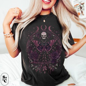 K&ouml;nnte beinhalten: Schwarzes T-Shirt mit einem grafischen Druck eines Sch&auml;dels, eines Nachtfalters und Rosen. Die Grafik ist von einem Kreis mit drei Mondsicheln und dem Wort "DOC" in der Mitte umgeben.