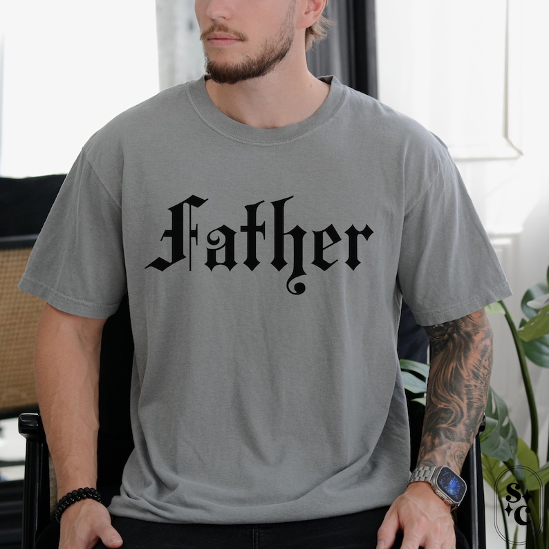 May include: A gray t-shirt with a black gothic font design that reads "Father".