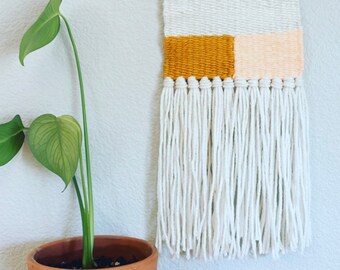 Puede incluir: Un tapiz de pared tejido blanco con un diseño geométrico amarillo y melocotón. El tapiz tiene un borde de flecos y está sujeto a una varilla de madera.