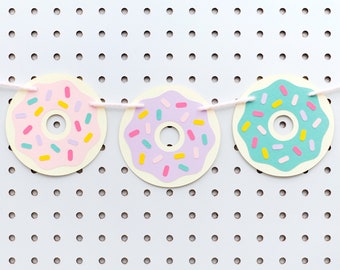 May include: A donut garland with five paper donuts in pink, yellow, purple, blue, and green with white sprinkles. The donuts are strung together with white string and are hanging on a white pegboard.