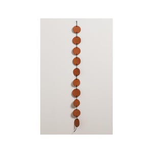 Puede incluir: Un adorno de pared decorativo con una serie de piezas circulares de cerámica de color terracota conectadas por un cordón negro. Los círculos están espaciados uniformemente y suspendidos de un gancho de metal, creando un diseño vertical. El fondo es una pared blanca lisa.