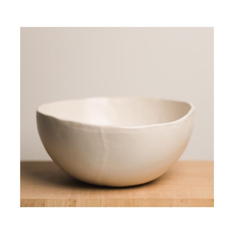 May include: A large, white ceramic bowl with a slightly irregular rim. The bowl is sitting on a wooden surface.