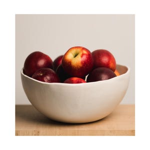 May include: A white ceramic bowl filled with red apples. The bowl has a slightly wavy rim and is sitting on a wooden surface.