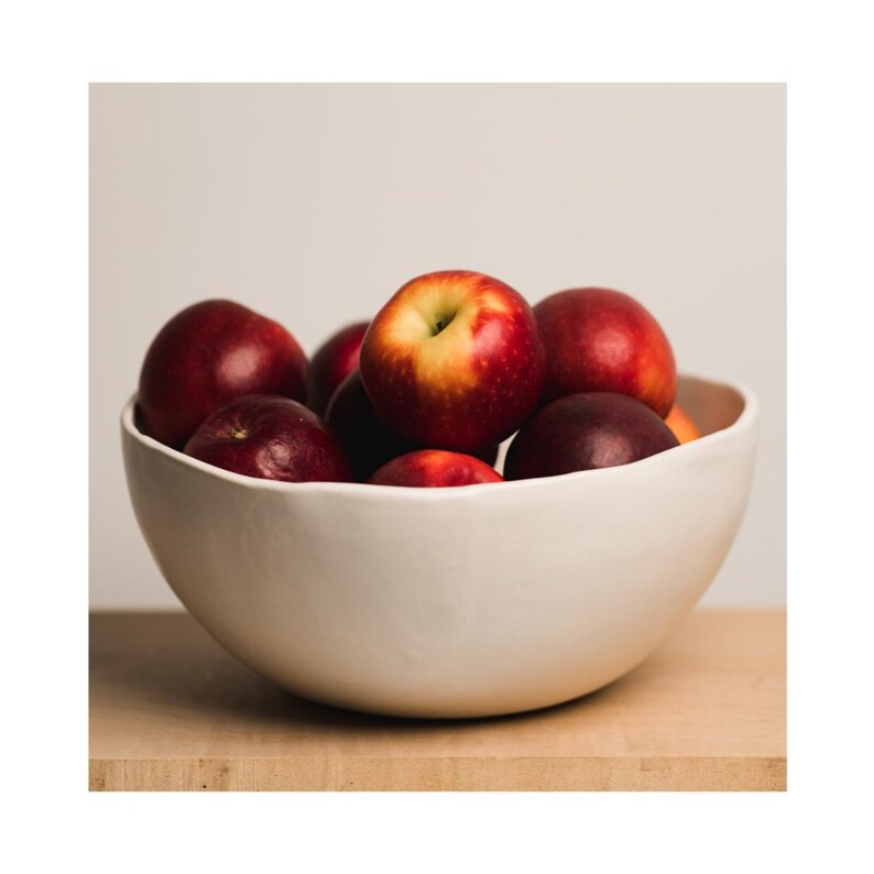 May include: A white ceramic bowl filled with red apples. The bowl has a slightly wavy rim and is sitting on a wooden surface.
