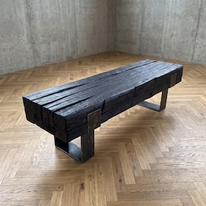 Op de afbeelding: Zwarte houten bank met een rechthoekige bovenkant en metalen poten. De bank is gemaakt van dik, verweerd hout en heeft een modern, industrieel ontwerp. De bank is ongeveer 152 cm lang.