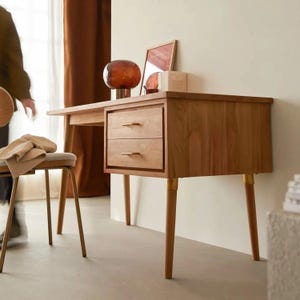May include: A wooden desk in a mid-century modern style, featuring two drawers with brass handles. The desk has a light brown finish and is supported by four tapered legs. A chair with a beige cushion is nearby. A decorative lamp and framed art are on the desk.