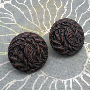 May include: Two antique bronze-colored buttons with intricate leaf and paisley patterns. The buttons are round and have a raised design. They are set against a floral patterned background.