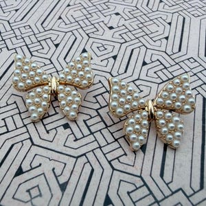May include: Two butterfly-shaped decorative pins featuring a gold-toned metal frame filled with small, round, white faux pearls arranged in a grid pattern across the wings. Each butterfly has a central gold body connecting the two wing sections.