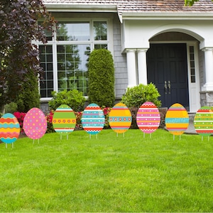 May include: Eight colorful Easter egg yard signs with different patterns. The signs are arranged in a row on a green lawn in front of a house.