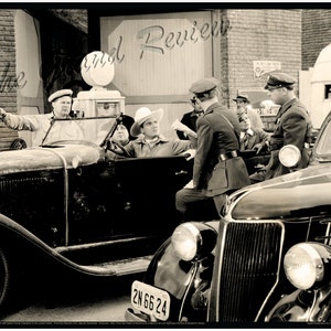 May include: Sepia-toned image of a vintage scene with classic cars and people in period attire. A group of men, some in uniforms, are gathered near the cars. One man in a car points to the left. The scene evokes a sense of nostalgia.