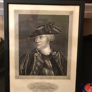 May include: A black and white portrait of a man in a military uniform wearing a tri-cornered hat. The portrait is framed in a black frame and has a decorative border with stars. The name "PAOLI" is written in a decorative script below the portrait.