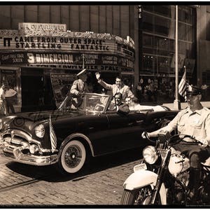 May include: A black and white photo of a vintage car with a group of people inside. The car is being escorted by a police officer on a motorcycle. The car is driving past a movie theater marquee that reads "It Came From Outer Space" and "Startling Fantastic".