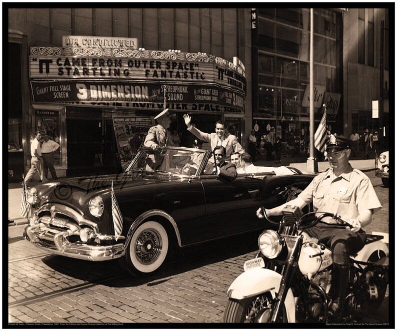 May include: A black and white photo of a vintage car with a group of people inside. The car is being escorted by a police officer on a motorcycle. The car is driving past a movie theater marquee that reads "It Came From Outer Space" and "Startling Fantastic".