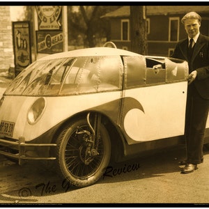 May include: Sepia-toned image of a streamlined, vintage car with a unique, enclosed design. The car has a white body with a wave-like design and a riveted metal roof. A man in a suit stands next to the car. The car has a license plate that reads "RP 58 39".